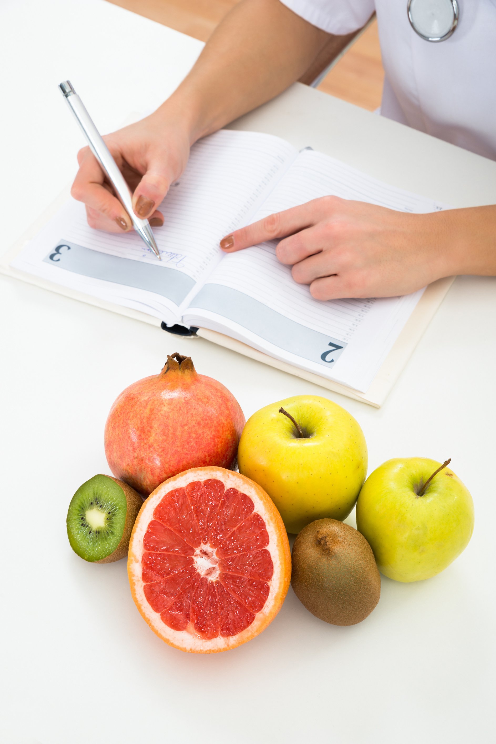 Dietician Writing Prescription With Fruits On Desk